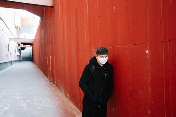 a young man in black sportswear and a baseball cap with a medical mask and backpack on his face near the red wall.