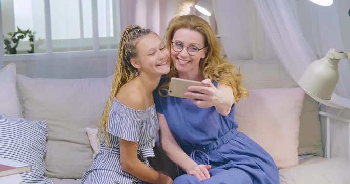 Happy positive mother with grown-up adult daughter making selfie using smartphone. Make funny face expressions, stick out tongue. Mom and child enjoying spending time together