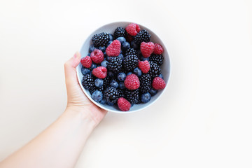 Female hand holding a white bowl with a mix of fresh summer berries - blackberry, blueberry,...