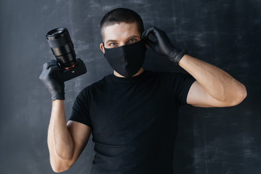 Young man Photographer in a protective black mask and gloves on a dark background holds a camera in his hands taking pictures. Concept Photo business and coronovirus.
