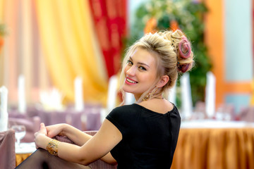 A beautiful, young blonde with a fashionable hairstyle and model appearance, sitting in a restaurant on a chair, laughing joyfully looking at the camera.