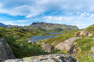 Mountain landscape with a rocky hill and green meadow overlooking a tranquil lake, surrounded by rugged peaks and blue sky in Norway.