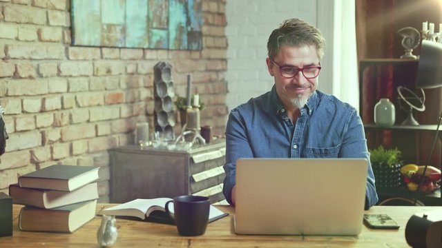 Older man tele working with laptop computer at home office in living room, thinking, looking busy.