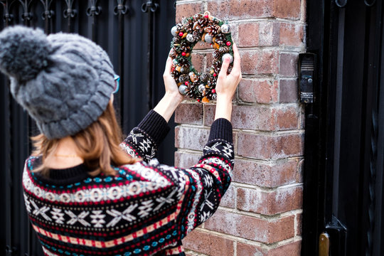 The pretty girl wearing cozy sweater with ornament is hanging up colorful handmade Christmas wreath with cones and glistening balls on the door outside