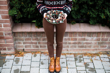 The pretty girl wearing cozy sweater with ornament is holding colorful handmade Christmas wreath with cones and glistening balls in her hands