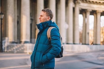 A young blond man in a jacket and with a backpack stands in the morning light on a deserted square in St. Petersburg against the background of the columns of the Kazan Cathedral