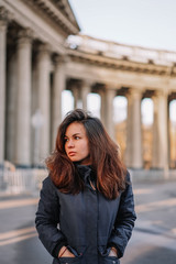 Obraz premium A young brunette girl with long hair in a jacket stands in the rays of the morning dawn on a deserted square in St. Petersburg against the background of the columns of the Kazan Cathedral