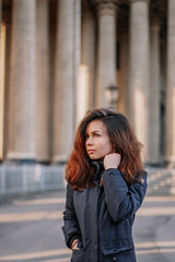 A young brunette girl with long hair in a jacket stands in the rays of the morning dawn on a deserted square in St. Petersburg against the background of the columns of the Kazan Cathedral