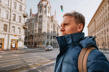 A young man in a jacket and a backpack stands at a traffic light against the background of an...