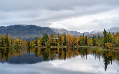 Lake reflecting autumn forest at Marsliden, Sweden, with calm water and mountains under a cloudy sky