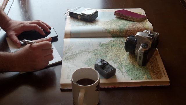 Detail of a man's hands writing in a notebook while preparing a trip with a map and photographic equipment on a table. Concept: preparing a trip