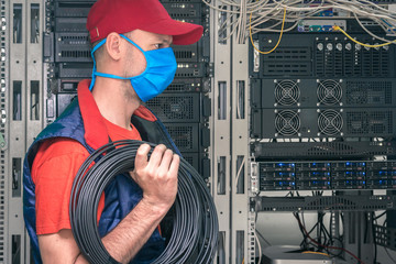 A technician in a medical mask works in the server room. A specialist with a coil of black fiber...
