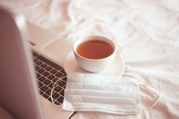 Fresh cup of tea with medical mask staying on open laptop in bed. Quarantine time. Good morning. Social distancing.