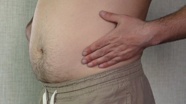 A chubby man stands by the wall. Stroking his stomach with his hands. Excess weight. Obesity. A fragment of the body. Without a face.