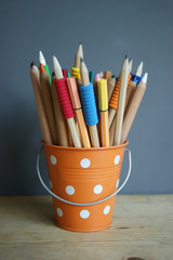 A Set Of multi colored Pencils and pens in An orange dotted Toy Bucket against a gray background