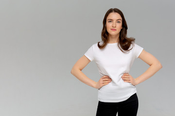 Beautiful smiling young woman in white t-shirt is holding hands on hip and looking at camera. Three quarter length studio shot on monochromatic background.
