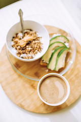Idea for healthy and hearty breakfast. Avocado toast, homemade granola with peanut butter, cup of coffee on wooden tray. 