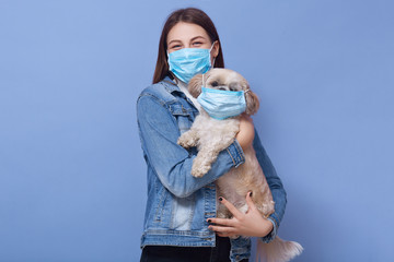 Indoor shot of woman dresses denim jacket protective flumask holds dog pet in medical mask....