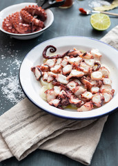 Closeup view of a delicious dish of octopus slices on a potato bed. At the back there are a dish with octopus arms, half a lime and a spoon full of paprika.