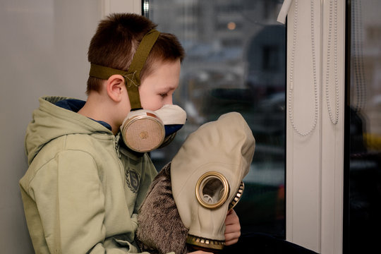 a child in a respirator and jacket holds a toy in a gas mask and sits on the windowsill next to a Maine Coon cat.
