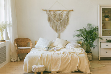 Modern scandi boho style bedroom interior with decorative pillows, green plant and diy macrame wall panel. Light warm cozy comfortable home. jomo, weekend breakfast in bed. Slow living concept.