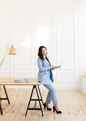 Fototapeta na wymiar A serious business working freelance girl a woman with black hair in a pale blue suit working on a laptop and phone at a wooden Desk against a white wall in the home office