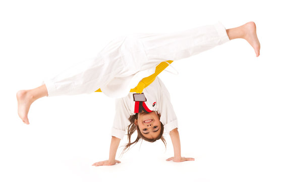 Little girl in white kimono with yellow belt, smiling and standing on her arms, stretching her legs into twine