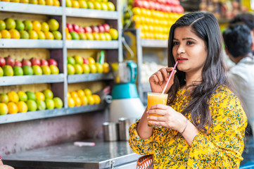 Young happy smiling Indian woman drinking orange juice outdoor in the market with colorful background of fruits.