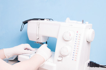 a woman in white disposable medical gloves sews on a sewing machine a protective face mask made of gray cotton fabric, blue background, we sit at home concept, quarantine 2020, shortage of masks
