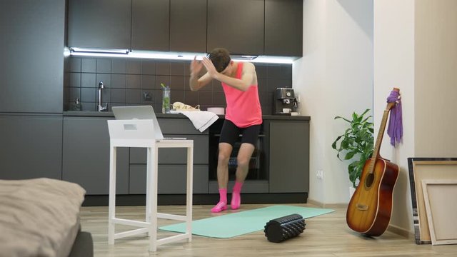 Young guy doing aerobic and fitness exercises at home using online training app on laptop computer. Funny man with mustache and pink top from 80's working out in kitchen staying home during quarantine