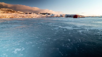 Low angle timelapse sliding left to right along surface of frozen lake at sunset.  Ice fishing shack on ice packed up part way through.  Golden clouds and mountains in background dipping into shadow. - Powered by Adobe