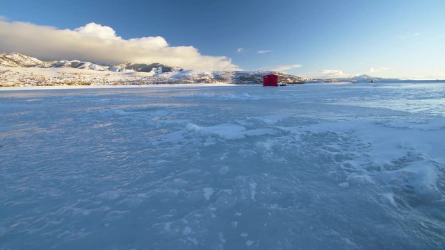 Low angle camera slider pulling back and right to left along the surface of frozen over lake at golden hour.  Ice fishing shack in background on ice.  Clouds over golden mountains in distance.