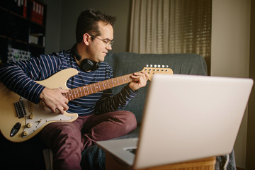 Boy taking an online course to learn to play the electric guitar. He is sitting on the sofa at...