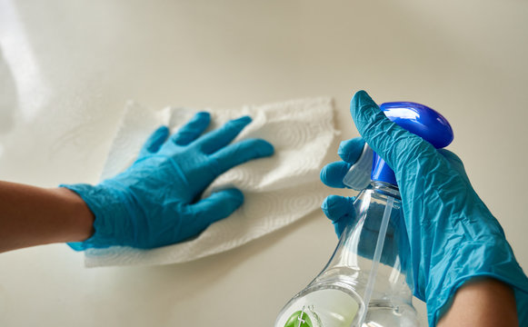 Human Hands Wearing Blue Disposable Latex Glove Cleaning The White Surface With A White Paper Towel Holding A Clear Plastic Bottle