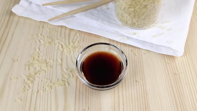 Soy sauce in transparent glass gravy boat. Traditional ingredient of asian cuisine. Wooden table, close-up, scattered grains of rice, chopsticks, white napkin