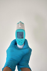 Human hand with blue disposable glove hold a digital infrared thermometer for body temperature scan...