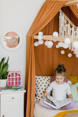  A little girl takes a book from a shelf Lies in a bed which stands in a stylish children's room and reads a fairy tale.
The room has a very beautiful interior design