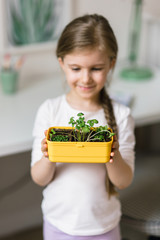 6 years old girl holding sprouted seedlings in her hands
She stands in her children's room by the window and smiles. She did the homework of biology in quarantine.