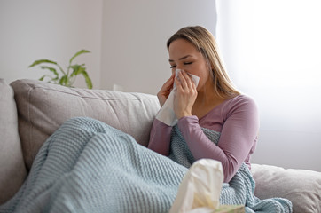 Sick woman blowing nose, sitting under the blanket. Sick woman with seasonal infections, flu, allergy lying in bed. Sick woman covered with a blanket lying in bed with high fever and a flu, resting.