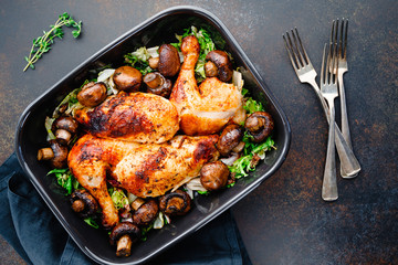 Top view of roasted chicken legs and breasts with fresh salad and mushrooms in black dish.