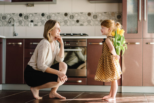 Cute little girl greeting mother and gives her a bouquet of flowers tulips on kitchen. Mother's day concept. Mom and daughter smiling. Happy family holiday and togetherness. Side view.