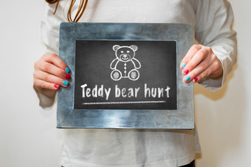 Girl holding a chalkboard with bear drawing and the text Teddy bear hunt. Teddy bear hunts keep children entertained during the corona virus lockdown.