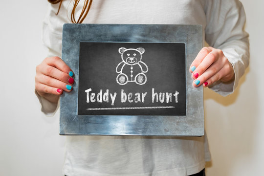 Girl Holding A Chalkboard With Bear Drawing And The Text Teddy Bear Hunt. Teddy Bear Hunts Keep Children Entertained During The Corona Virus Lockdown.