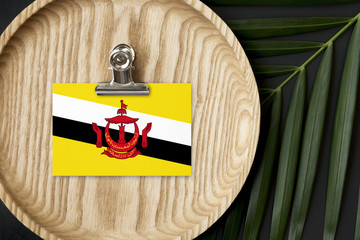 Brunei flag tagged on wooden plate. Tropical palm leaves monstera on background. Minimal national concept.