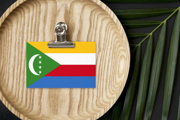 Comoros flag tagged on wooden plate. Tropical palm leaves monstera on background. Minimal national...