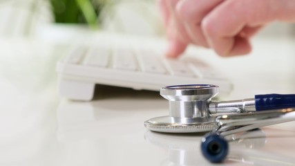 Doctor working at the workplace. Clinic worker typing on laptop. Stethoscope on foreground. Close up.