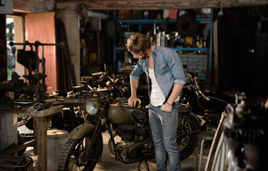 A young man with a hipster look works in his workshop repairing vintage motorcycles.