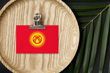 Kyrgyzstan flag tagged on wooden plate. Tropical palm leaves monstera on background. Minimal...