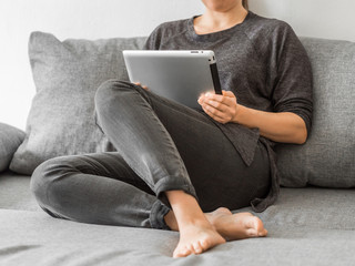 Young girl watching lessons on a tablet sitting on gray sofa. Business woman on quarantine. Home office, online education, remote work, distance learning, stay home concept