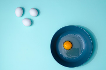 Three white eggs and a blue plate with broken egg on the table on the mint blue background